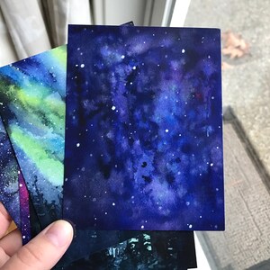 May include: Three watercolor paintings of the night sky with stars and galaxies. The paintings are on white paper and are in shades of blue, purple, and green.