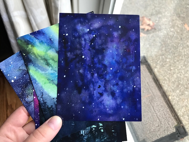 May include: Three watercolor paintings of the night sky with stars and galaxies. The paintings are on white paper and are in shades of blue, purple, and green.