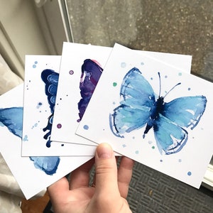 May include: Four watercolor paintings of butterflies on white card stock. The butterflies are painted in shades of blue and purple with white accents. The paintings are all different sizes and shapes.