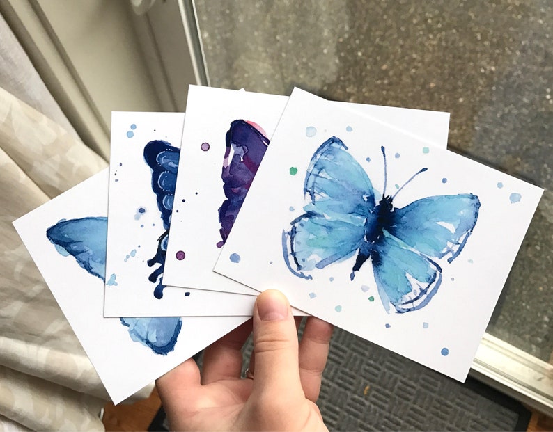 May include: Four watercolor paintings of butterflies on white card stock. The butterflies are painted in shades of blue and purple with white accents. The paintings are all different sizes and shapes.