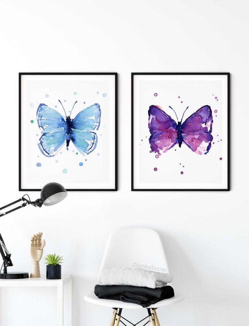 May include: Two framed watercolor paintings of butterflies. The butterfly on the left is blue and the butterfly on the right is purple. Both butterflies are on a white background with watercolor splatters.