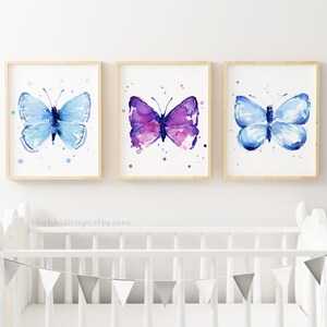 May include: Three framed watercolor paintings of butterflies. The butterflies are painted in shades of blue and purple with white accents. The paintings are displayed on a white wall above a white crib.