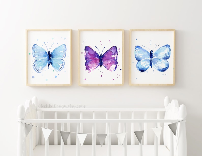May include: Three framed watercolor paintings of butterflies. The butterflies are painted in shades of blue and purple with white accents. The paintings are displayed on a white wall above a white crib.