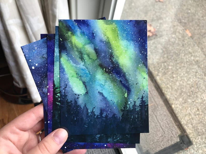 May include: A set of four watercolor paintings depicting the aurora borealis over a dark forest. The paintings are on thick paper and have a textured surface. The colors are vibrant and the details are intricate.