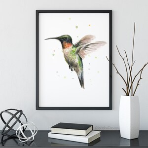 May include: A watercolor painting of a hummingbird in flight with a black frame. The hummingbird is green, red, and brown with a long beak. The background is white with small green, yellow, and orange dots.