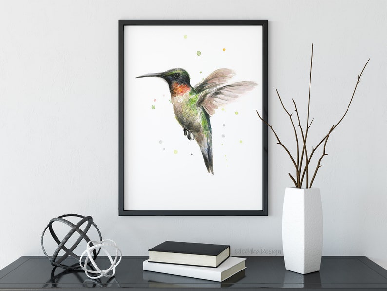 May include: A watercolor painting of a hummingbird in flight with a black frame. The hummingbird is green, red, and brown with a long beak. The background is white with small green, yellow, and orange dots.