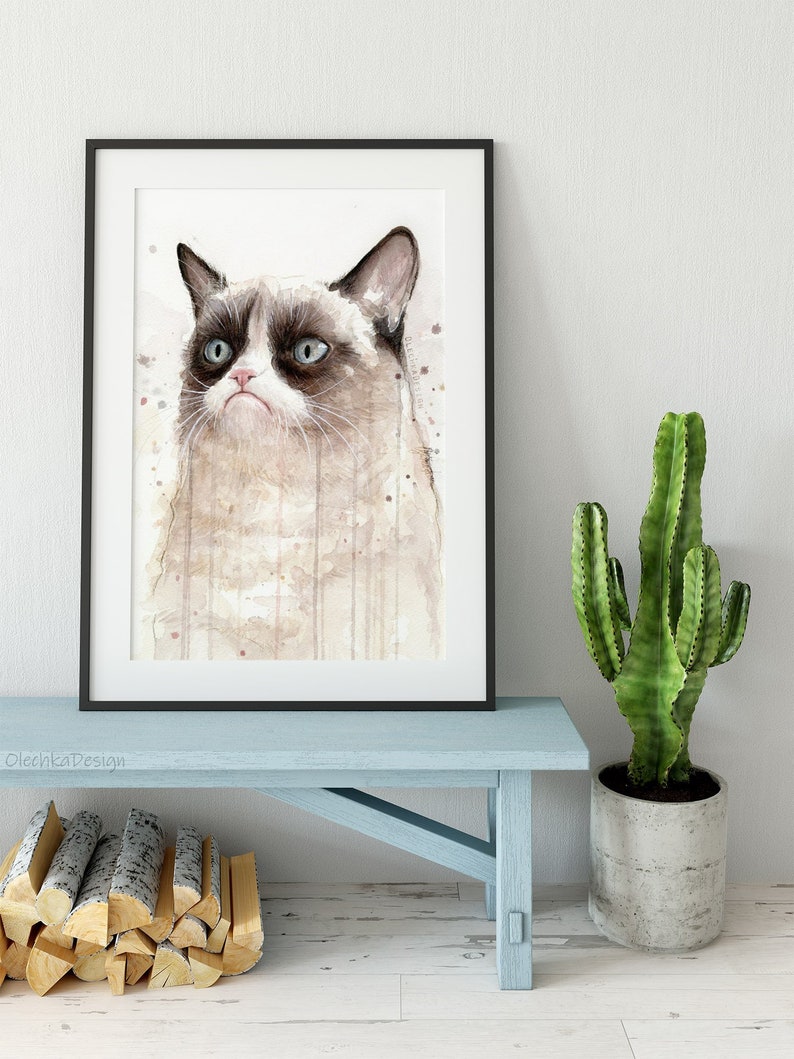 May include: A watercolor painting of a grumpy cat with a white and gray coat. The cat is looking to the right with a serious expression. The painting is framed in a black frame and is hanging on a white wall.