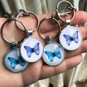 May include: Four silver keychains with round glass charms featuring watercolor butterfly designs. The butterflies are in shades of blue and purple.