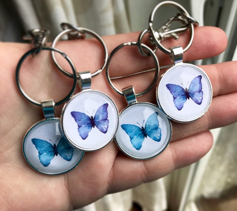 May include: Four silver keychains with round glass charms featuring watercolor butterfly designs. The butterflies are in shades of blue and purple.