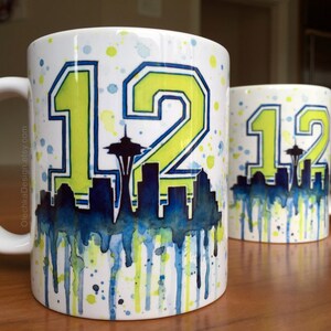 May include: Two white ceramic mugs with a watercolor design. The mugs feature a green and yellow number "12" with a blue and white cityscape silhouette. The cityscape includes a depiction of the Space Needle.