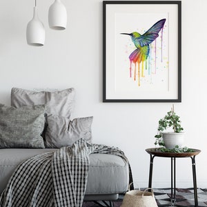 May include: A watercolor painting of a hummingbird in flight with colorful paint dripping down from its wings. The painting is framed in black and displayed on a white wall.