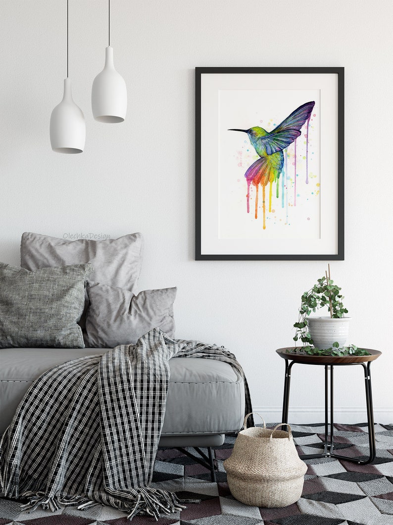 May include: A watercolor painting of a hummingbird in flight with colorful paint dripping down from its wings. The painting is framed in black and displayed on a white wall.