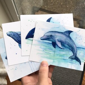 May include: Set of three watercolor paintings featuring marine animals. The paintings depict a humpback whale, an orca, and a dolphin. The paintings are on white card stock with blue and green watercolor washes.