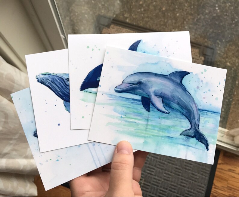 May include: Set of three watercolor paintings featuring marine animals. The paintings depict a humpback whale, an orca, and a dolphin. The paintings are on white card stock with blue and green watercolor washes.