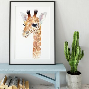 May include: A watercolor painting of a baby giraffe with brown and white spots. The giraffe is looking to the left and has a light brown nose and large, dark eyes. The painting is framed in a black frame and is hanging on a white wall.