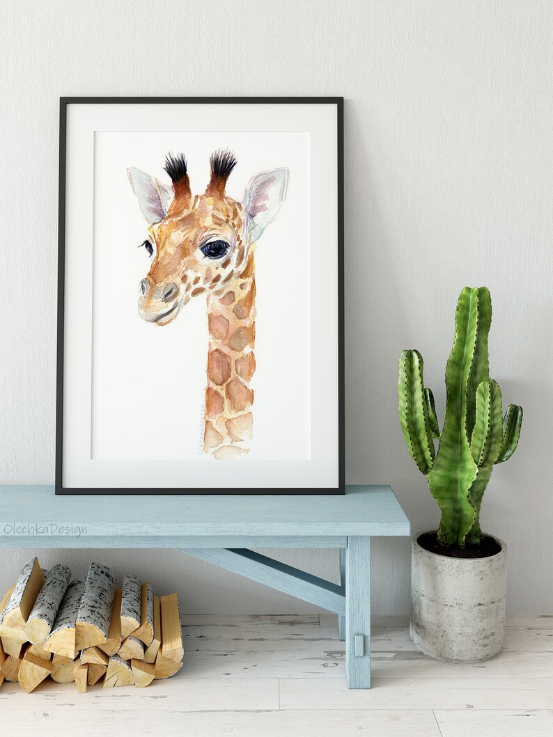 May include: A watercolor painting of a baby giraffe with brown and white spots. The giraffe is looking to the left and has a light brown nose and large, dark eyes. The painting is framed in a black frame and is hanging on a white wall.