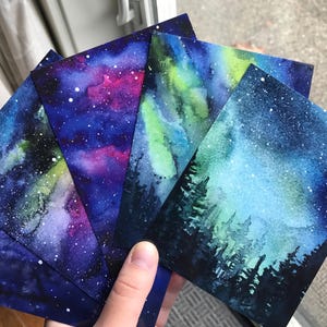 May include: Five watercolor paintings of a starry night sky with a galaxy of stars and nebulae. The paintings are on paper and have a blue, purple, and green color scheme.