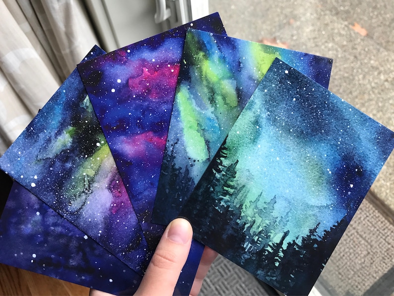 May include: Five watercolor paintings of a starry night sky with a galaxy of stars and nebulae. The paintings are on paper and have a blue, purple, and green color scheme.