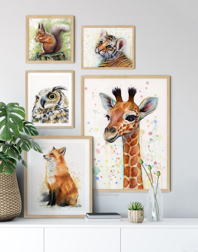 May include: A set of five framed watercolor paintings featuring various animals. The paintings include a squirrel, a tiger cub, an owl, a giraffe, and a fox. The paintings are all in a light wood frame and are arranged on a white wall.