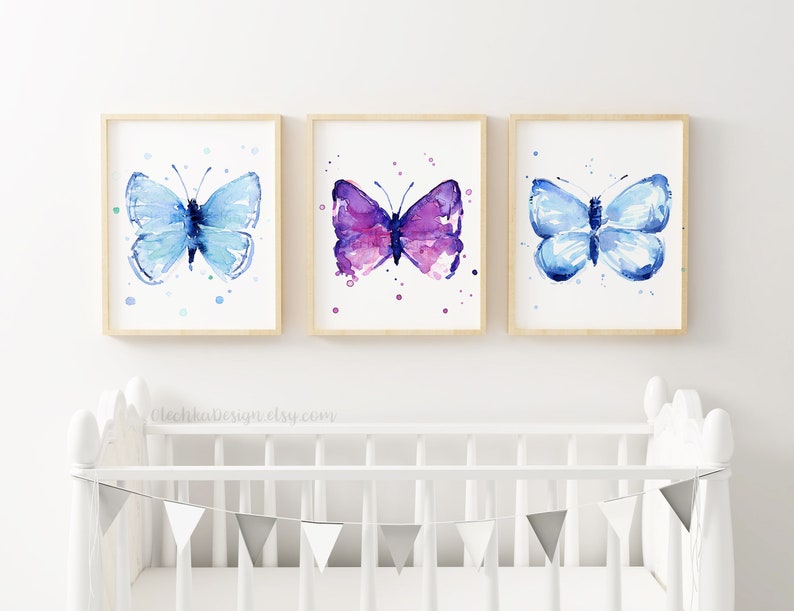 May include: Three framed watercolor butterfly prints. The butterflies are in shades of blue and purple, with a light wooden frame. The artwork is displayed above a white crib with a gray and white banner.