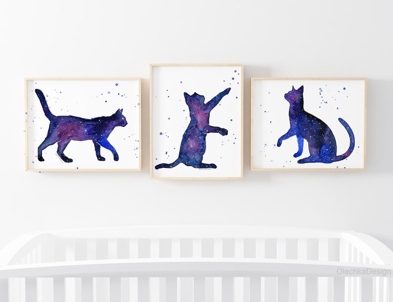 May include: Three watercolor prints of galaxy cat silhouettes. The cats are in different poses, one walking, one sitting, and one standing on its hind legs. The background is white with a few watercolor splatters.