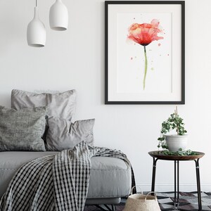 May include: A watercolor painting of a single red poppy flower with green stem and leaves. The painting is framed in a black frame and hanging on a white wall above a gray sofa with pillows and a black and white throw blanket.