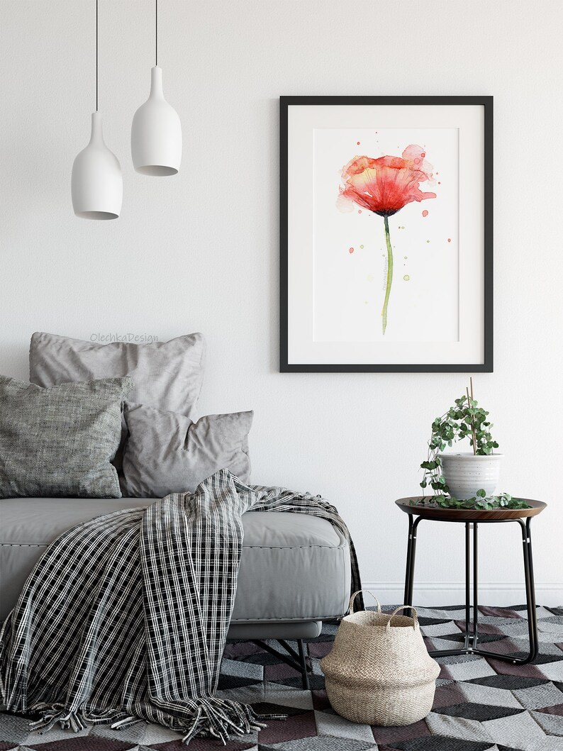 May include: A watercolor painting of a single red poppy flower with green stem and leaves. The painting is framed in a black frame and hanging on a white wall above a gray sofa with pillows and a black and white throw blanket.