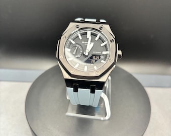May include: A G-Shock Casio wristwatch featuring an octagonal black metal bezel with eight visible screws, a dark gray analog-digital dial with white markers, and a light blue silicone strap. The digital display shows 10.25 and 19.