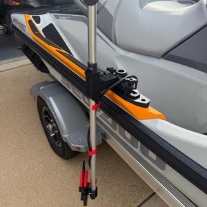 May include: A silver metal pole mounted to the side of a white and orange personal watercraft. The pole features a black top cap, black mounting hardware, and two bright red clips securing a black cable running down its length to a black base.