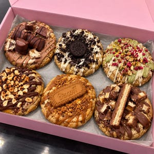 May include: A pink cardboard box containing six large round cookies with various toppings. The cookies feature chocolate bars, crushed sandwich cookies, chopped nuts, dried berries, Biscoff biscuits, and caramel drizzles on a light-coloured dough base.