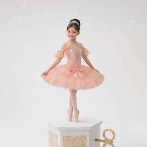 May include: A young girl in a pink ballet tutu and pointe shoes stands on a white music box. The tutu has a layered design, and the bodice is embellished with gold accents. A decorative key is attached to the side of the music box.