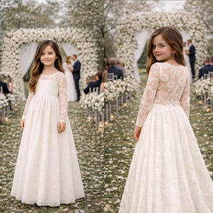 May include: A young girl in a long, ivory lace dress with long sleeves, standing in a garden setting. The dress features a round neckline and a full skirt. The background includes a floral arch and wedding guests.