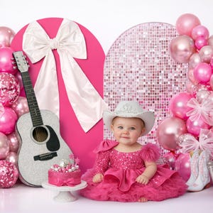 May include: A baby in a pink dress and cowboy hat sits in front of a backdrop with pink balloons, a guitar, and a cake. The backdrop includes a large pink heart shape with a white bow and a mosaic wall.