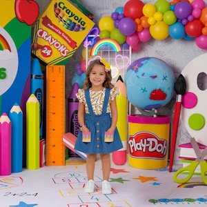 May include: A young girl stands in front of a colorful backdrop with school supplies. The scene includes large crayons, pencils, a ruler, a globe, and a Play-Doh container. A rainbow and the number 2026 are also visible.