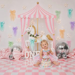 May include: A carousel-themed first birthday scene with a smiling child. The backdrop features a pink and white striped canopy, a white unicorn carousel, and decorative bows. A cake with a carousel design sits in front of the child.