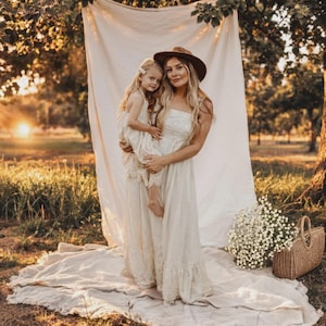 May include: A woman and a young girl in matching cream-colored dresses pose outdoors. The woman wears a brown hat. A white sheet hangs in the background. A basket of white flowers sits nearby.