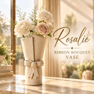 Op de afbeelding: Een crèmekleurige keramische vaas met een gespikkelde afwerking, gevormd als een ingepakt boeket met een strik. De vaas bevat witte pioenen en een lichtroze roos. De tekst rechts luidt: Rosalie RIBBON BOUQUET VASE.