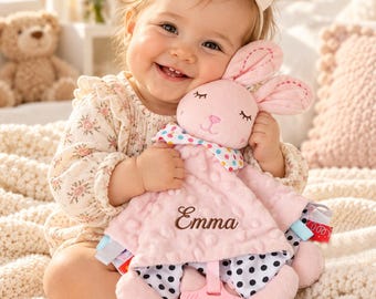 May include: A soft pink plush security blanket featuring a stuffed rabbit head with embroidered closed eyes and a nose. The blanket has a dimpled texture, a colorful polka dot collar, and the name Emma embroidered in brown script.