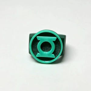 May include: A dark green ring featuring a circular face with a raised, bright mint green Green Lantern logo emblem. The emblem consists of a central circle surrounded by a ring, with two horizontal bars extending from the sides of the inner circle.