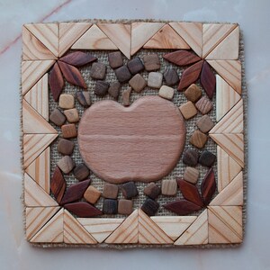 Puede incluir: Un posavasos de madera con forma de manzana con un acabado de madera marrón. El posavasos está rodeado de un mosaico de pequeños cuadrados y triángulos de madera en tonos marrones, rojos y beige. El mosaico está colocado sobre un fondo de arpillera.