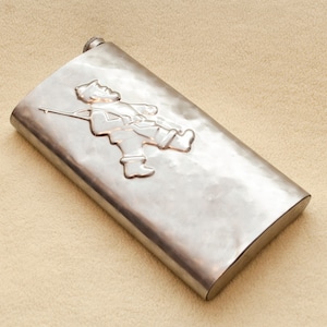 May include: A silver metal hip flask with a raised design of a person carrying a rifle. The flask has a rounded shape and a smooth surface.