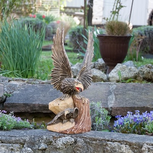 Könnte beinhalten: Eine Holzschnitzerei eines Adlers mit ausgebreiteten Flügeln. Der Adler sitzt auf einem Baumstumpf, an dessen Basis ein kleinerer Vogel geschnitzt ist. Die Schnitzerei ist in Braun- und Schwarztönen bemalt.