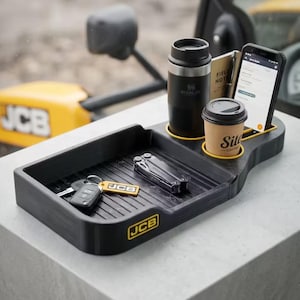Puede incluir: Una bandeja organizadora de plástico negro con el logotipo amarillo de JCB, diseñada con una base acanalada. Incluye dos portavasos circulares con un vaso Stanley negro y un vaso de café de papel, además de una ranura para un teléfono y una libreta.
