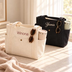 May include: Two canvas tote bags, one cream and one black, each featuring a front pocket with a snap button, side pockets holding a tumbler, and a pair of sunglasses hanging from a strap. The cream bag is embroidered with Winona, the black with Yasmin.