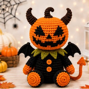 May include: A crocheted stuffed toy featuring an orange pumpkin head with a black stitched jack-o'-lantern face, dark brown horns, and a small stem. The body is black with two orange buttons, green collar, black bat wings, and an orange devil tail.