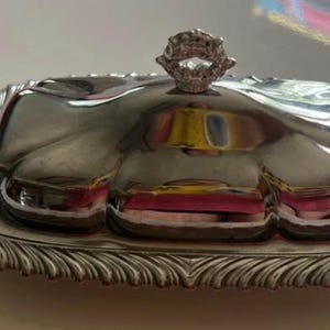 May include: A rectangular silver-toned butter dish with a polished, reflective domed lid and a decorative, scalloped edge base. The lid features a small, ornate handle at the top center, and the entire piece has a smooth, metallic finish.