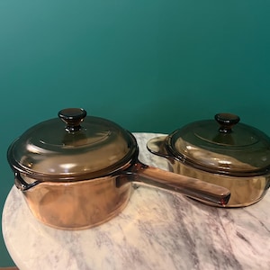 May include: Two pieces of amber-colored transparent glass cookware. One piece is a saucepan with a long handle and a matching lid. The second piece is a round casserole dish with two side handles and a matching lid. Both items are made of glass.