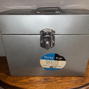 May include: A silver metal Porta-file document box with a top carrying handle and a central locking latch. A circular sticker on the front reads Porta-file for home or office, durable baked enamel finish, easy carry balanced handle, and holds over 500 documents.