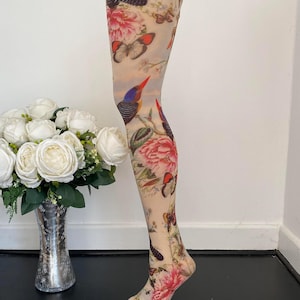 Peut inclure: Collants beiges transparents ornés d'un imprimé multicolore représentant divers papillons, oiseaux et grandes fleurs roses. Le vêtement est présenté sur un mannequin en forme de jambe, mettant en valeur le motif botanique et animalier détaillé.