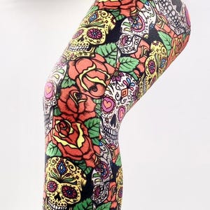 May include: Leggings featuring a vibrant, all-over print of colourful sugar skulls and red roses on a black background. The pattern includes intricate decorative details on the skulls, green leaves, and various floral elements covering the fabric.